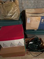 Stacked plastic totes and cardboard boxes including one with Christmas tree print, holding multiple string lights and accessories.