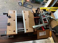 Black & Decker Workmate portable workbench with wooden top and integrated adjustable clamps, metal frame with signs of wear, various clamps and a wooden handled tool placed on top and inside a box.