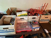 Overview of the lot showing boxes of hardware items, extension cords, and tool sets on a table.