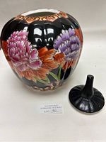 Black ceramic vase with hand painted purple and pink flowers, gold accents, and removable lid with pointed handle, photographed from a side angle.