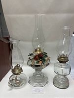 Three glass oil lamps of varying heights, one decorated with holiday pinecone arrangement.