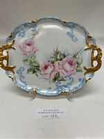 Front view of Limoges porcelain plate showing hand-painted pink roses, blue scroll details, and gold trim on handles and edges.