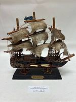 Front view of scale model ship showing fabric sails, wooden and metal elements, mounted on a labeled black wooden base.