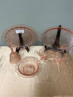 Two oval pink depression glass platters displayed on black stands, along with a hand bell, cream and sugar set, and small bowl arranged together on a surface.