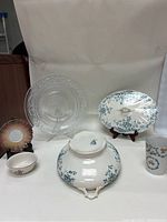 View of soup tureen with lid, clear depression glass plate, Austrian floral mug, small saucer and gold rim bowl arranged together.