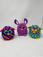 Three colorful Furbies, two with eye glasses-like coverings and one with large upright ears, against white background
