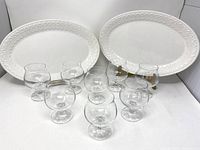Eight clear crystal brandy snifters arranged in a group in front of two white ceramic oval platters with embossed borders.