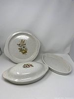 Three cream speckled stoneware pieces: round platter with floral design, covered oval baker, and open oval baker, displayed for condition and design detail.
