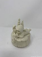 Close-up photo of the Dept 56 Snowbabies music box showing side view of child figure lying on snow-covered tree stump with bird and two star-topped trees.