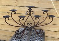 Front view showing full 7-candle metal candelabra with symmetrical scrollwork design, placed on outdoor wooden surface against a vertical wood fence.