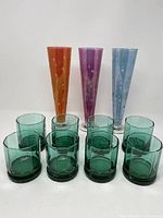 Full set of eight green octagonal old fashioned glasses arranged in front of three tall colored pilsner glasses in orange/pink, purple, and blue.