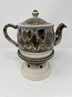 Front view of the lava drip glaze vintage Beauceware teapot sitting on its warming stand, showing the textured dark brown and black drip glaze pattern with handle and spout.