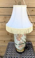 Full view of lamp with white fabric lampshade and visible floral ceramic base on wood table against wood wall.