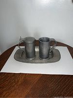 Front view showing two small cylindrical pewter tankards and an oval tray on a wooden table.