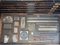 Top view of various small to large printing plates including brass, steel, and wood blocks in a wooden tray