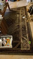 Full shot of rectangular olive green area rug with brown and beige geometric and floral patterns, partially covered by household items.