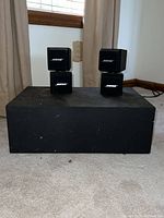 Four Bose satellite cube speakers stacked and placed on the large black subwoofer enclosure in room with carpet and window.