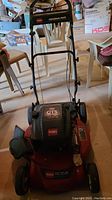 Front view of the red Toro lawn mower with black engine cover showing the model and 'Personal Pace' on handlebar. Lawn mower positioned indoors with visible surrounding chairs and boxes.