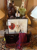 Photo showing dark wood magazine rack with carved edges, Buddha wooden figurine, ceramic panda figurine, six vintage Saki glasses with floral patterns on a black tray, green ceramic vase, and Asian style framed art piece with visible broken glass.