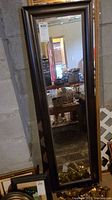 Tall mirror with dark wood frame leaning against concrete wall, showing clear reflective surface and beaded border detail.