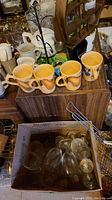Four hand-painted ceramic mugs on wooden box with metal mug tree and white teapot in background, box with vintage clear glasses below