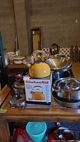 Photo showing a yellow KitchenAid kettle on its box surrounded by stainless steel pots and bowls and coated frying pans on a wooden countertop.