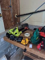 Wide view of the collection of garden tools arranged on a wooden rack in a garage corner, showing Ryobi leaf blower, green trimmer, red handheld trimmer, battery chargers, and extension cords.