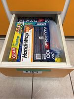 Drawer opened to show collection of wrapping products including Alcan aluminum foil, Kirkland cling wrap, Handi-Wrap plastic wrap, Glad cling wrap, and ziplock bags.