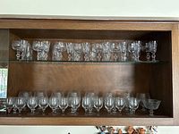 Crystal stemware and glasses arranged on two wooden shelves with glass shelf inserts. Top shelves contain various snifters and cut crystal glasses. Bottom shelves hold plain lowball glasses and small bowls.