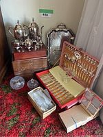 Photo showing silver tea and coffee set on wooden boxes with crystal and glass pieces around, two red cutlery boxes open with silver and gold cutlery inside, empty wooden box underneath.
