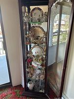 Full view of fine china and crystal items in a tall dark wood and glass display cabinet with mirrored back.