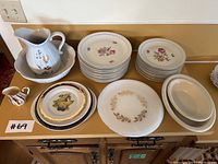 Wide view of all china items laid out on kitchen counter, including large pitcher and basin, stacks of plates of various sizes, and small creamers.