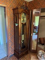Full frontal view of tallcase clock showing carved wooden crest, glass door, base and surrounding decor