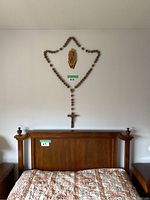 Wide shot showing the large wooden rosary hanging on a wall above a queen-sized bed with part of the bed and nightstands visible.