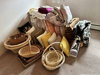 Wide view showing large collection of pillows, folded quilts, blankets, and assortment of wicker baskets stacked on carpeted floor near window.
