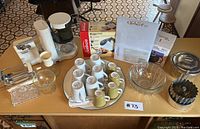 Wide view showing Braun coffee maker, mugs, glass bowls, metal bundt pan, plastic storage container and other kitchen items on wooden table.