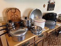 View of the cookware lot on wooden kitchen cabinet, including stainless steel pots and pans with glass lids, Presto pressure cooker, white enamel roasting pan, wooden cutting boards, and textured roasting pans.