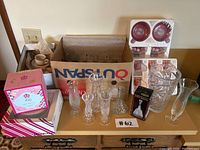 Overview of the glassware and crystal lot showing boxed and unpackaged items on a wooden piece of furniture against a wall.