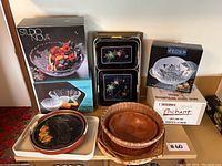 Photo showing multiple boxes and trays stacked including Studio Nova glass bowl box, Peony glass dish box, black lacquer floral trays, and woven and wooden bowls.
