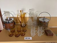 Full view of all items including vintage ice buckets, amber and clear glass decanters, matching glassware, and small boxed item.