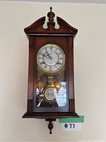 Full front view of antique wooden wall clock hanging on white wall, showing clock face with Roman numerals and pendulum behind glass.