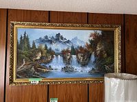 Front view of large landscape painting with mountains and waterfalls in ornate gold frame against wood paneled wall with partial view of lamps and furniture