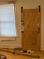 Full view of the wooden barn door leaning against a wall next to a window, with boxes and packaging visible on floor beneath.