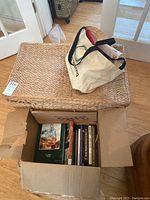 Photo showing a large rectangular straw woven trunk with a cream canvas tote bag placed on top. Next to the trunk is an open cardboard box filled with books and magazines.