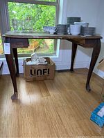 Full view of vintage wood desk showing curved legs and wooden top with some discoloration. Missing center drawer and visible damage to front feet.