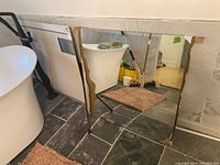 Photo shows two square frameless wall mirrors leaning against tiled wall and floor in a bathroom setting. Mirrors have visible damage on edges and corners, particularly the top left corner.
