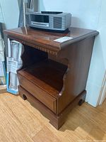 Angle view of wooden nightstand showing drawer with inside handle and open lower shelf. Some scratches and wear visible.