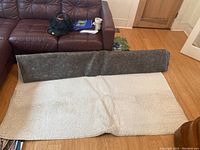 Ivory shag rug partially unrolled showing gray underside with visible damage, placed on wood flooring with brown leather couch in background