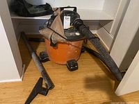 Orange Ridgid 9-gallon shop vacuum cleaner with black hose and black extension tubes on the floor, stored on wooden floor inside shelving area.