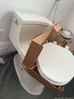 Photo showing the Koehler low flow toilet with toilet seat attached and extra toilet lid remaining in cardboard box on top of the seat.